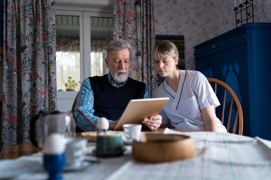 Female caregiver sitting beside senior patient solving crossword puzzle in tablet at home visit