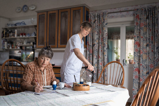 Female caregiver pouring coffee in cup for woman solving puzzle in newspaper at home