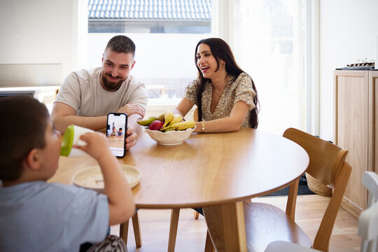 Happy parents showing photographs to son on smart phone while sitting at table in home