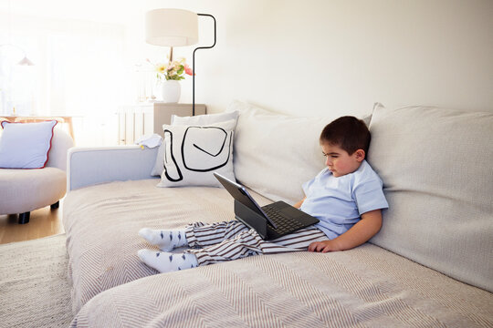 Cute boy sitting with laptop on sofa in living room at home