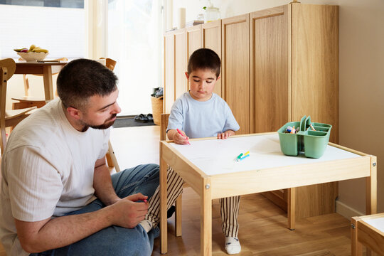 Father sitting beside boy coloring on paper at table in home