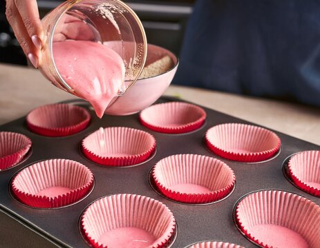 cropped hand pouring pink batter from measuring cup into cupcake liners