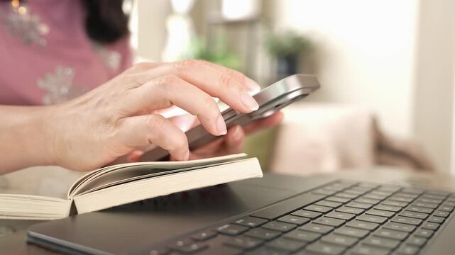 Woman using smartphone to research information while studying at home, scrolling typing beside laptop and book, showing technology, communication, adult learning, work and thoughtful decision making
