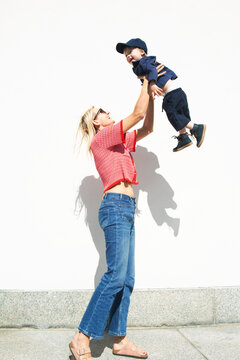 Full length of happy mother picking up son while standing near wall at sunny street