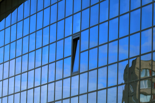 Close-up of a modern glass building facade featuring a grid of rectangular panes; a single tilted window breaks the pattern, revealing a glimpse of interior; vivid blue sky and soft clouds reflect acr