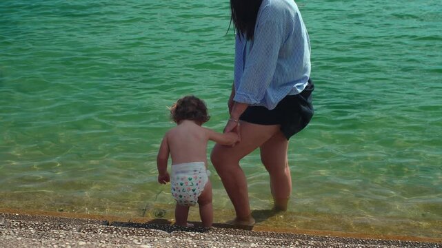 Mother guiding toddler into shallow lake water while holding hand during summer outing capturing trust, safety and authentic childhood exploration outdoors
