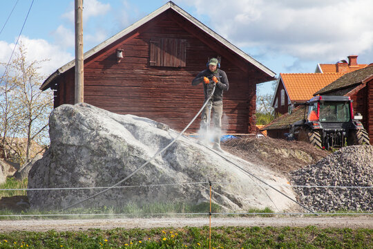 Mature man in protective gear operating pneumatic rock drill on large boulder against cloudy sky