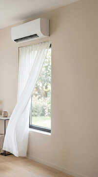 Modern air conditioner installed above window in bright room. Interior wall with blowing white curtain. Climate control system for home comfort and summer cooling during hot weather season.
