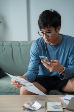 Asian man sitting on sofa holding smartphone and checking bill statement document while planning personal monthly financial expense at home