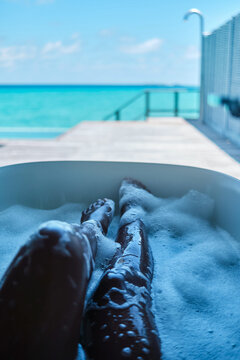 A white person soaks legs in bubble bath with ocean view