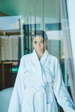 A woman in a white bathrobe looks pensively through a glass window