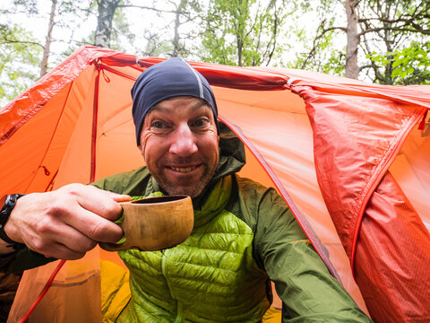 Smiling male traveler holding wooden cup while sitting in camp tent