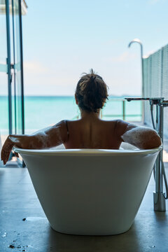 A white woman enjoys a bubble bath in a modern tub with an ocean view
