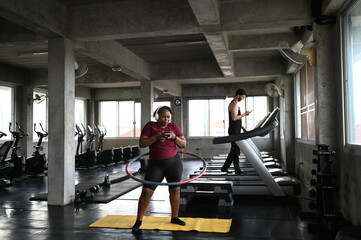 Dedicated plus-size black woman using a smartphone while exercising with a weighted hula hoop on a yellow mat inside a modern concrete gym facility during a daily fitness workout routine. © Kanlaya
