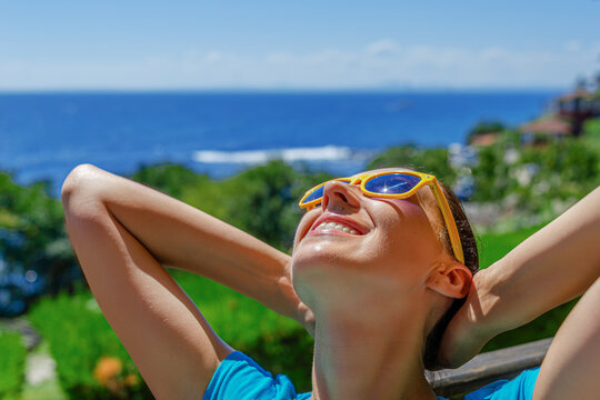 A smiling woman in yellow sunglasses relaxes outdoors with her hands behind her head, enjoying the sunny weather near a tropical ocean view.