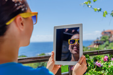 Fototapeta premium A young woman wearing sunglasses holds up a digital tablet that reflects her smiling face on a sunny balcony overlooking the sea.