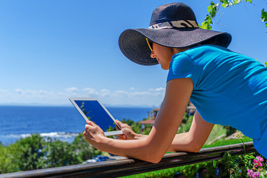 A woman wearing a hat holds a digital tablet while leaning on a balcony railing overlooking the sea.