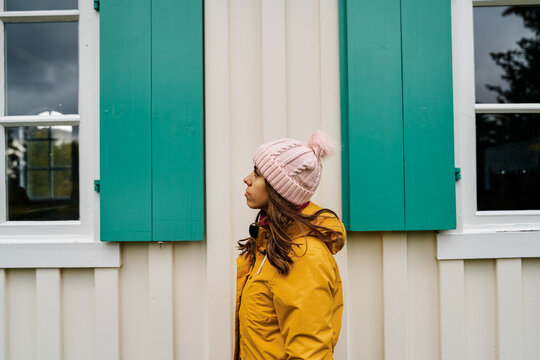 White Woman in yellow jacket and pink hat standing by a building