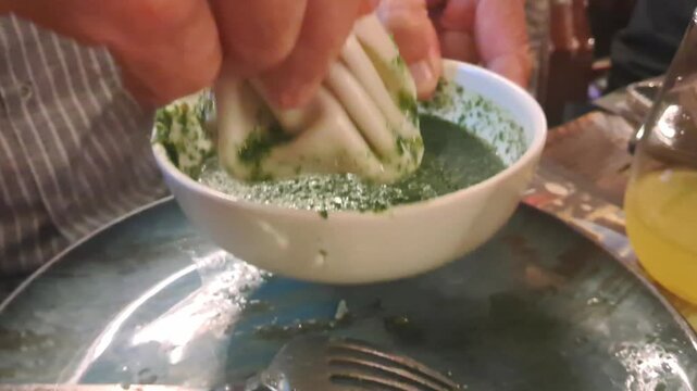 A close-up view of a person holding a Georgian khinkali dumpling by its top knot and dipping it into a bowl of green herb sauce