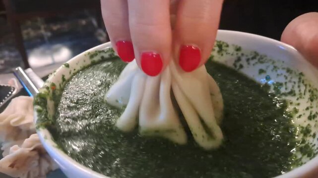 A close-up view of a person holding a Georgian khinkali dumpling by its top knot and dipping it into a bowl of green herb sauce