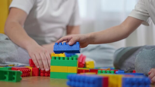 Blocks on floor, girl adds yellow part while kids build. Boy selects blocks as siblings cooperate. Tower of blocks rises while kids adjust pieces. Girl supports tower with both hands on blocks.