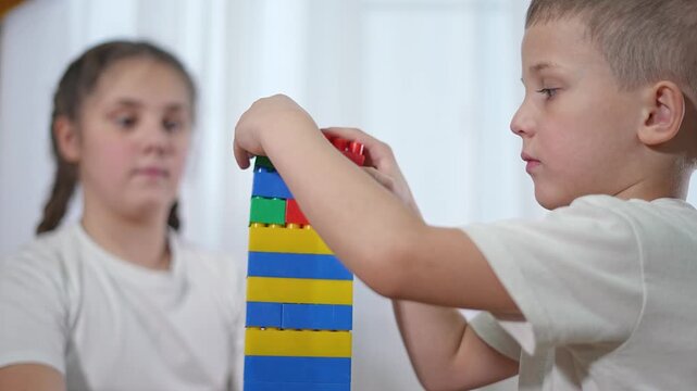 Boy smiles completing block tower. Kids celebrate fun building process. Girl observes creative result pride. Siblings playing blocks on floor. Creative kids build tall structure using favorite blocks