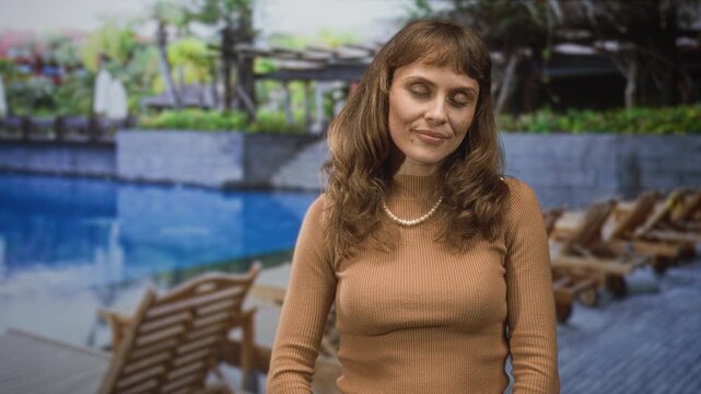 Woman resting head on hands with closed eyes beside a pool and lounge chairs at a building; serenity relaxation.
