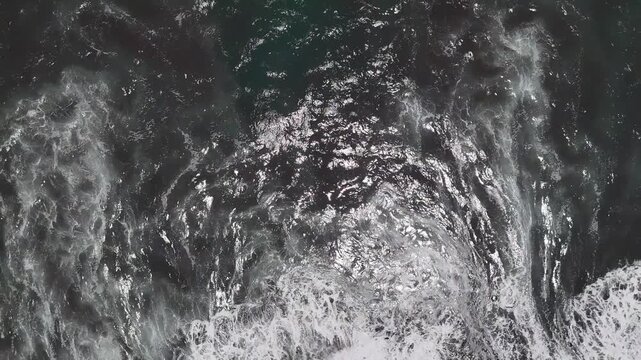 Drone ascends above an ocean wave breaking onto a black volcanic sand beach, revealing white sea foam patterns and dark Pacific coastal water from above.