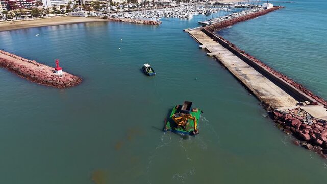 Aerial view of a dredging barge with yellow excavator and tugboat operating near a harbor entrance with red lighthouse, stone breakwater, and marina with boats in background