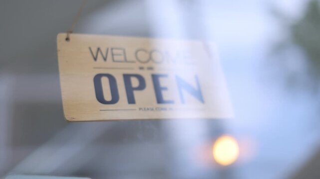 A wooden Welcome Open sign hanging on a glass door of a shop, filmed with a soft-focus background.