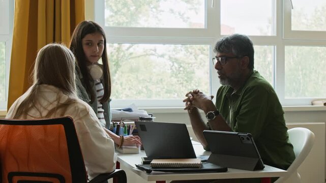 Middle aged South Asian male teacher explaining lesson to two Caucasian teenage girls sitting at desk with laptop. Perfect for educational marketing, tutoring services, or school mentorship promos