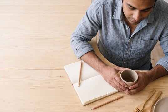 Thoughtful Indian man sitting at a wooden table with an open blank notebook and holding a ceramic cup