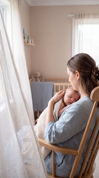 Caring Mother Sitting in Wooden Rocking Chair Holding Her Sleeping Newborn Baby in a Sunlit Nursery Room