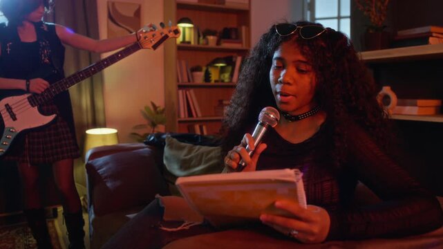 African American young adult female holding microphone and reading lyrics from notebook while singing during band rehearsal. Ideal for youth lifestyle, music education, and hobby campaigns