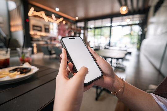 Person holds phone with blank screen, fingers gently gripping edges. Restaurant background softly blurred, warm lights glow overhead. Fingers show casual, relaxed grip, suggesting leisurely use