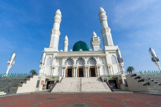 White mosque stands tall under bright blue sky. Green dome gleams above arched entrances and twin minarets. Broad stone steps lead to grand doors flanked by decorative arches