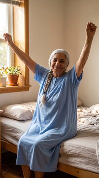 A happy senior woman with grey hair stretches her arms while sitting on her bed in the morning sunlight.
