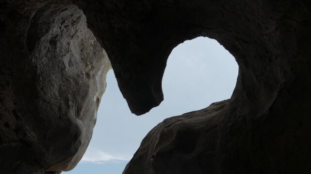 A static handheld shot looking through a jagged, winding opening in the rocky walls of the ancient cave city of Uplistsikhe, Georgia. The dark stone frames the bright sky outside.