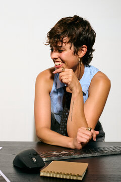 A woman with short hair smiles thoughtfully at her business office desk. She rests her chin on her hand, gazing at a laptop screen. Her expression conveys focused creativity and quiet satisfaction.  D