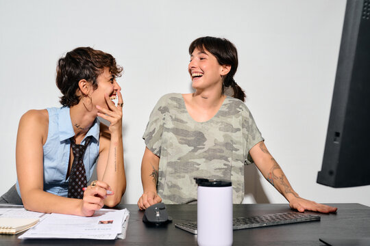 Two women share a joyful laugh at their desk. They&rsquo;re engaged in a lighthearted moment, hands near their faces as they smile.