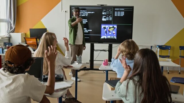 Teenagers sitting at desks raising hands during lesson with Indian middle-aged teacher pointing at interactive screen. Ideal for promoting modern STEM education and interactive learning
