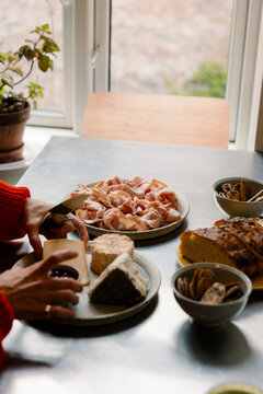 Woman assembling cured ham, cheese, and bread platter by sunny window