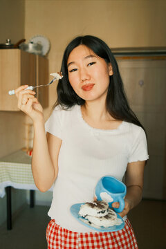 Young smiling asian woman eating cake and holding a mug at home 