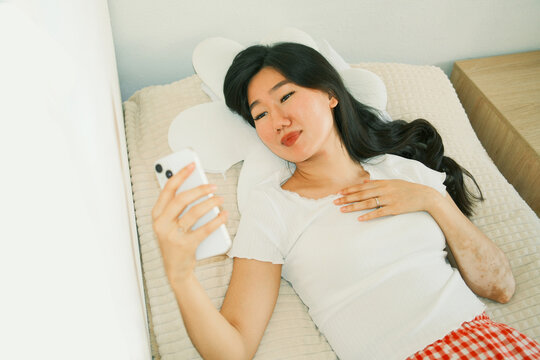 Overview of happy young asian woman using smartphone lying in bed