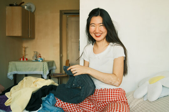 Happy young asian woman smiling, sitting on bed with clothes 