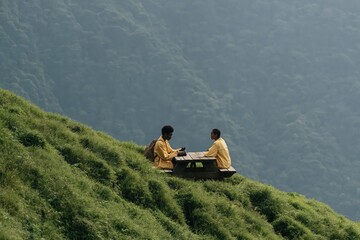 Fototapeta premium Two people sitting at picnic table on steep grassy hillside in matching yellow jackets for travel background and outdoor adventure banner