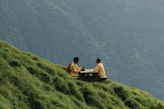 Two people sitting at picnic table on steep grassy hillside in matching yellow jackets for travel background and outdoor adventure banner