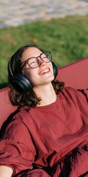 Smiling young woman wearing headphones outdoors relaxing on a bench in sunlight for banner background and portrait design