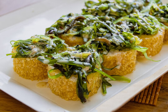 Crispy tempura sushi rolls topped with chuka seaweed salad and sesame nut sauce on white plate, Japanese fusion cuisine