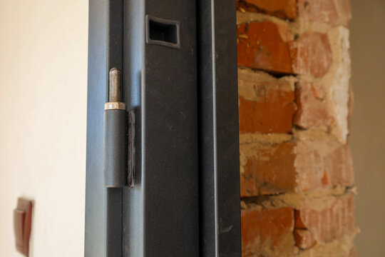 Close-up of a metal door hinge welded to the entrance door frame. The assembly is located next to an exposed red brick wall during installation and interior renovation. The hinge pin and rubber seal a